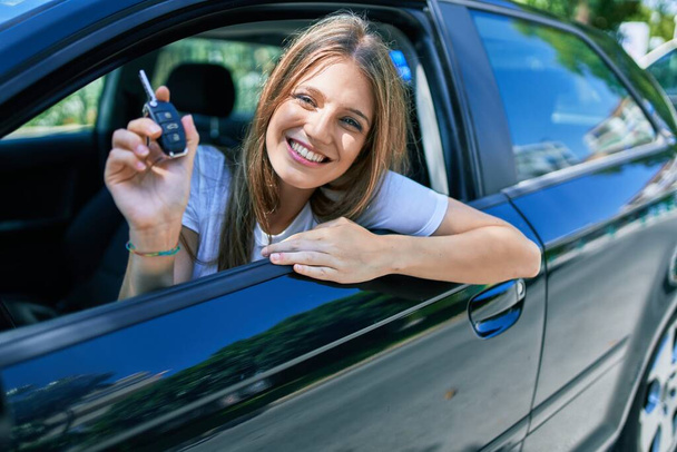 Junge schöne blonde Frau lächelt glücklich am Auto sitzend und zeigt Schlüssel. - Foto, Bild