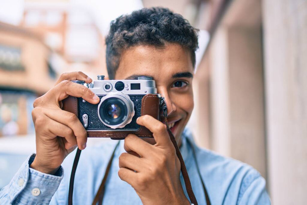 Joven turista latino sonriendo feliz usando la cámara vintage caminando por la ciudad. - Foto, Imagen