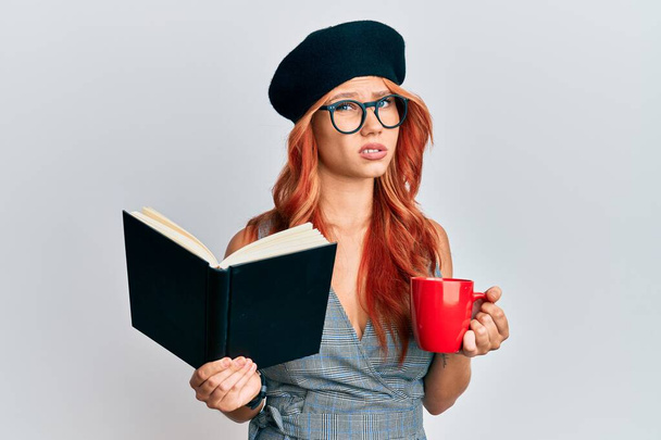 Młoda ruda kobieta w francuskim berecie czytająca książkę i pijąca kawę bez pojęcia i zdezorientowana ekspresja. koncepcja wątpliwości.  - Zdjęcie, obraz