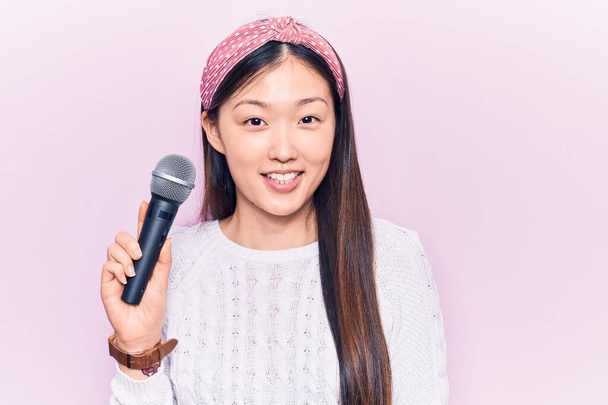 Νεαρή όμορφη Κινέζα γυναίκα τραγούδι τραγούδι χρησιμοποιώντας το μικρόφωνο αναζητούν θετικά και χαρούμενα στέκεται και χαμογελά με αυτοπεποίθηση χαμόγελο δείχνει τα δόντια  - Φωτογραφία, εικόνα