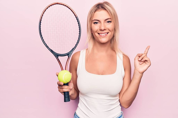 Νεαρή όμορφη ξανθιά αθλήτρια που παίζει τένις χρησιμοποιώντας ρακέτα και μπάλα πάνω από ροζ φόντο χαμογελώντας χαρούμενος δείχνοντας με το χέρι και το δάχτυλο στο πλάι - Φωτογραφία, εικόνα