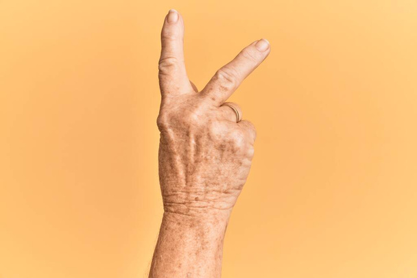 Mão caucasiana sênior sobre fundo amarelo isolado contando número 2 mostrando dois dedos, gesticulando vitória e símbolo vencedor  - Foto, Imagem