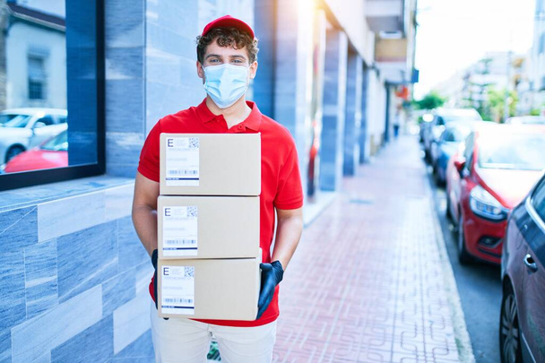 Young caucasian delivery man wearing coronavirus protection medical mask holding cardboard packages walking at city. - Фото, изображение