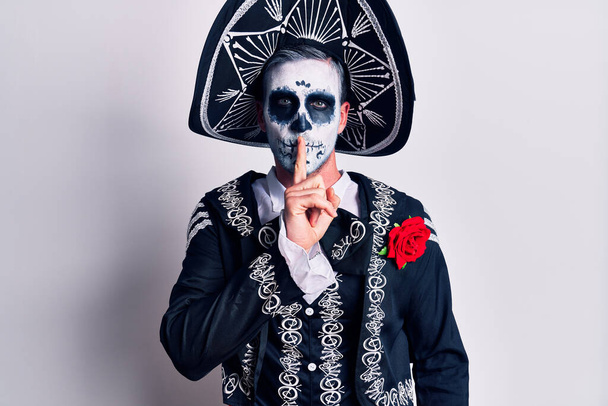 Молодой человек, одетый в мексиканский день мертвого костюма поверх белого, просит тишины с пальцем на губах. молчание и тайная концепция.  - Фото, изображение