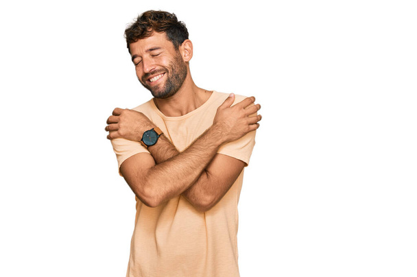 Knappe jongeman met baard die een casual tshirt draagt en zich vrolijk en positief knuffelt, vol zelfvertrouwen lachend. zelfliefde en zelfzorg  - Foto, afbeelding