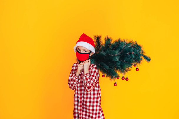 Child in front of a yellow background holding a Christmas tree. Christmas mood. She has a santa hat on her head - Photo, Image