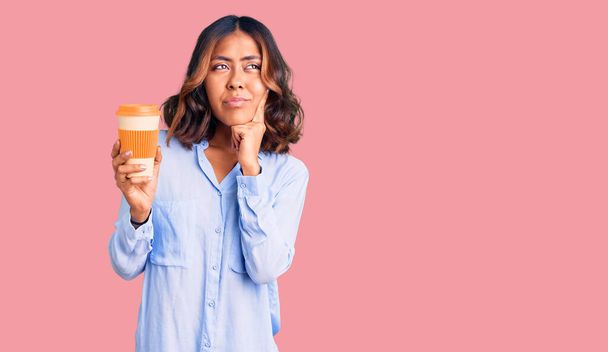 Молода красива змішана раса жінка, що п'є каву від забирання чашки серйозне обличчя, думаючи про питання з рукою на підборідді, вдумлива щодо заплутаної ідеї
  - Фото, зображення