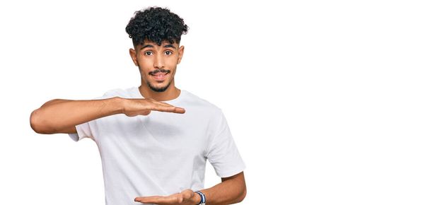 A fiatal arab férfi alkalmi fehér pólót visel gesztikuláló kezek mutatják nagy és nagy méretű jel, intézkedés szimbólum. mosolyogva néz a kamerába. mérési koncepció.  - Fotó, kép