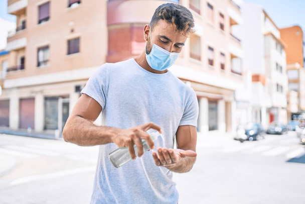 Jeune homme hispanique portant un masque médical à l'aide de gel snitizer main marche à la ville - Photo, image