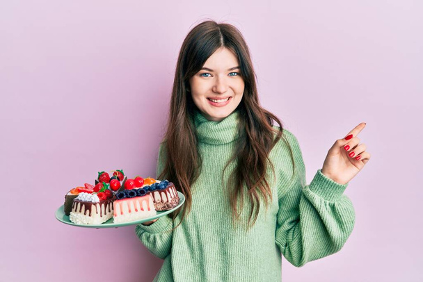 Young beautiful caucasian girl holding cake slices smiling happy pointing with hand and finger to the side  - Foto, imagen