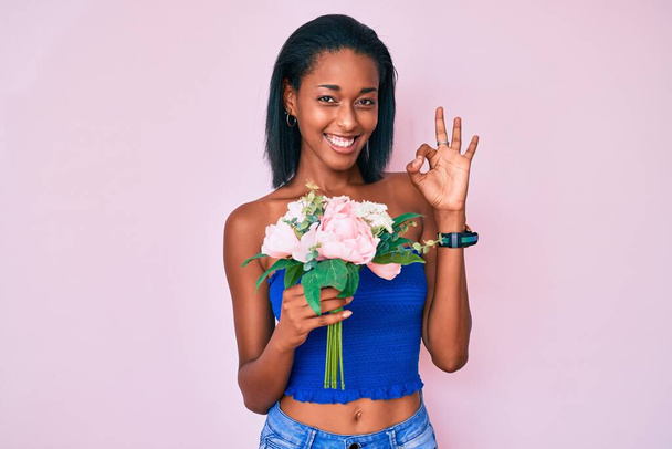 Junge afrikanisch-amerikanische Frau hält Blumen in der Hand und winkt mit den Fingern, lächelt freundlich und gestikuliert mit einem ausgezeichneten Symbol  - Foto, Bild