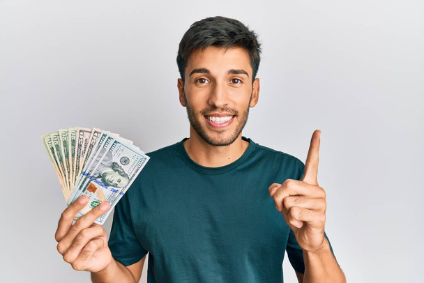 Joven hombre guapo sosteniendo dólares sonriendo con una idea o pregunta señalando el dedo con la cara feliz, número uno  - Foto, imagen