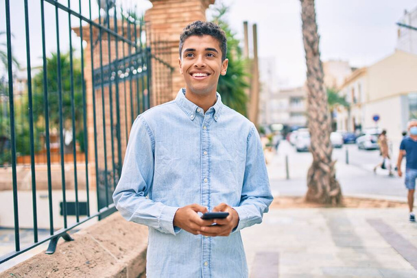 Молодий латинський чоловік посміхається щасливий, використовуючи смартфон, що йде по місту
. - Фото, зображення