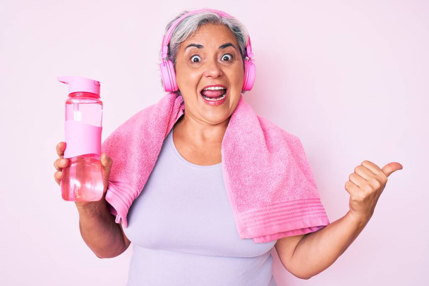 Mujer hispana mayor usando ropa de gimnasio y usando auriculares sosteniendo una botella de agua apuntando con el pulgar hacia el costado sonriendo feliz con la boca abierta  - Foto, imagen