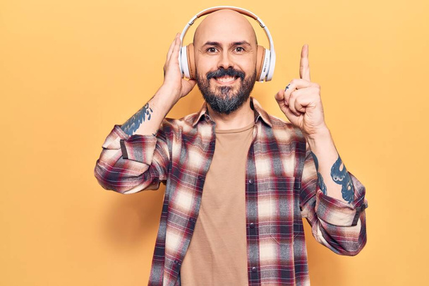 Jeune homme beau écoutant de la musique à l'aide d'écouteurs souriant avec une idée ou une question pointant du doigt avec un visage heureux, numéro un  - Photo, image
