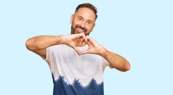 Handsome middle age man wearing casual tie dye tshirt smiling in love doing heart symbol shape with hands. romantic concept.  - Fotografie, Obrázek