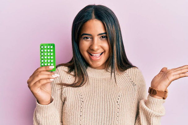 Молодая латиноамериканка с противозачаточными таблетками празднует достижение с счастливой улыбкой и выражением лица победителя с поднятой рукой  - Фото, изображение