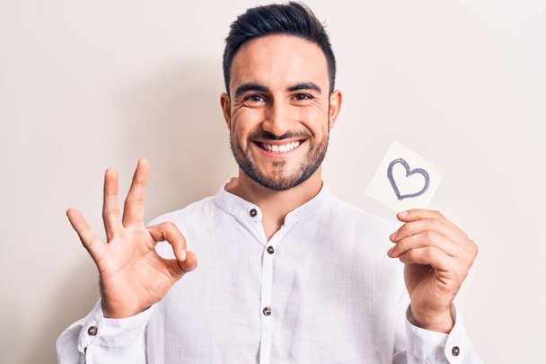 Young handsome romantic man with beard holding reminder with heart shape symbol doing ok sign with fingers, smiling friendly gesturing excellent symbol - Photo, Image