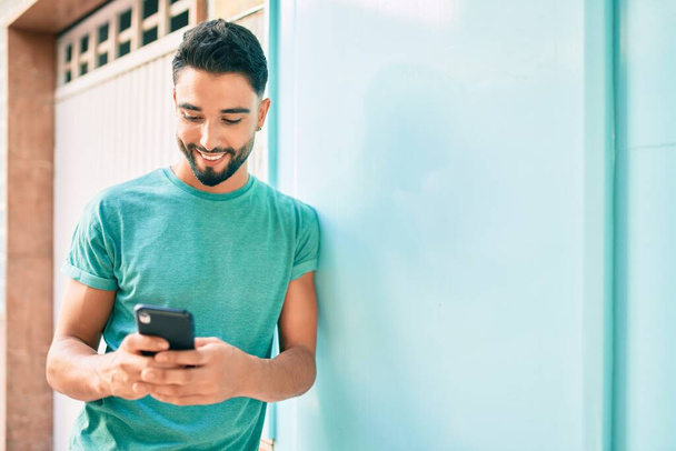 Jovem árabe homem sorrindo feliz usando smartphone na cidade. - Foto, Imagem
