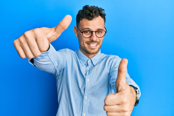 Jeune homme hispanique portant des vêtements décontractés et des lunettes approuvant faire un geste positif avec la main, pouces levés souriant et heureux pour le succès. geste gagnant.  - Photo, image