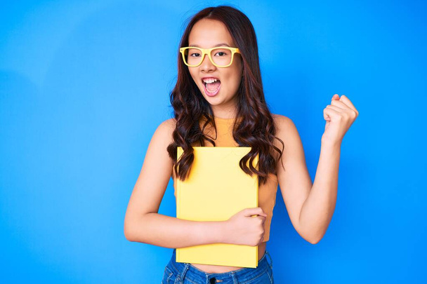 Young beautiful chinese girl wearing glasses holding book screaming proud, celebrating victory and success very excited with raised arms  - Photo, Image