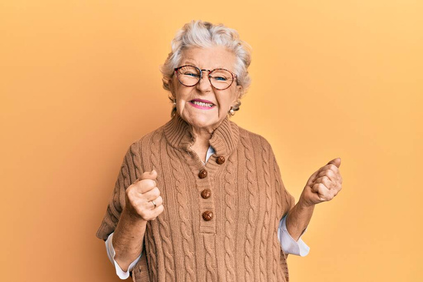 Ältere grauhaarige Frau in lässiger Kleidung und Brille, die vor Erfolg nur so strotzt, mit erhobenen Armen und geschlossenen Augen, die den Sieg lächelnd feiert. Siegerkonzept.  - Foto, Bild