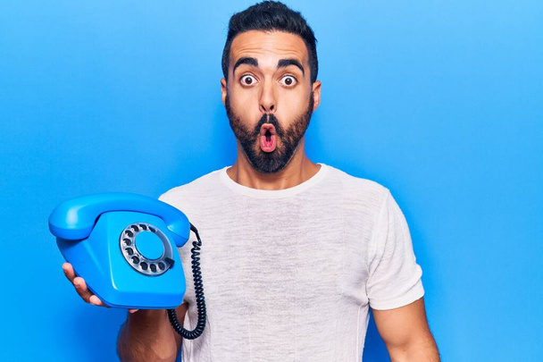 Mladý hispánec drží starý telefon vyděšený a ohromený s otevřenými ústy pro překvapení, nevěřící tvář  - Fotografie, Obrázek