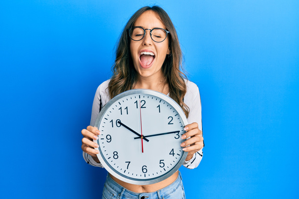 Mladá bruneta žena drží velké hodiny s úsměvem a hlasitě se smát, protože legrační šílený vtip.  - Fotografie, Obrázek