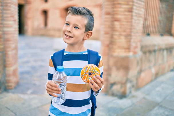 Adorable étudiant caucasien garçon souriant heureux tenant beignet et bouteille d'eau à la ville. - Photo, image