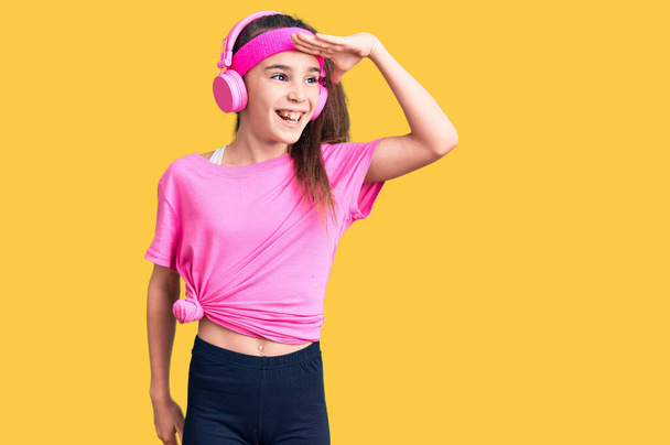 Χαριτωμένο ισπανόφωνο κορίτσι φορώντας ρούχα γυμναστικής και χρησιμοποιώντας ακουστικά πολύ χαρούμενος και χαμογελαστός κοιτάζοντας μακριά με το χέρι πάνω από το κεφάλι. Αναζήτηση έννοιας.  - Φωτογραφία, εικόνα