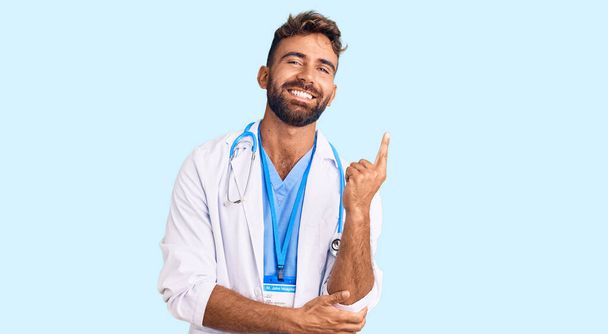Giovane uomo ispanico indossa uniforme medico e stetoscopio con un grande sorriso sul viso, puntando con mano e dito verso il lato guardando la fotocamera.  - Foto, immagini