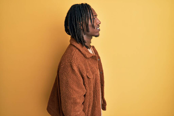 Afroamerikanischer Mann mit Zöpfen trägt braune Retro-Jacke, die zur Seite schaut, entspannte Profil-Pose mit natürlichem Gesicht und selbstbewusstem Lächeln.  - Foto, Bild