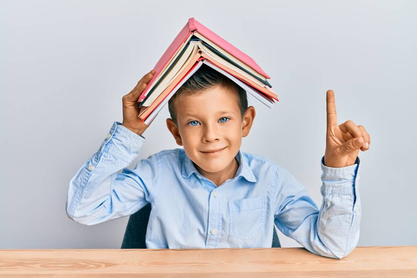 Очаровательный кавказский ребенок с книгой на голове, улыбающийся идеей или вопросом, указывающий пальцем со счастливым лицом, номер один  - Фото, изображение