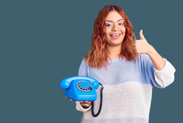 Jovem latina segurando telefone vintage sorrindo feliz e positivo, polegar para cima fazendo excelente e sinal de aprovação  - Foto, Imagem