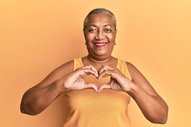 Mujer afroamericana mayor que usa estilo casual con camisa sin mangas sonriendo en amor haciendo forma de símbolo del corazón con las manos. concepto romántico.  - Foto, imagen