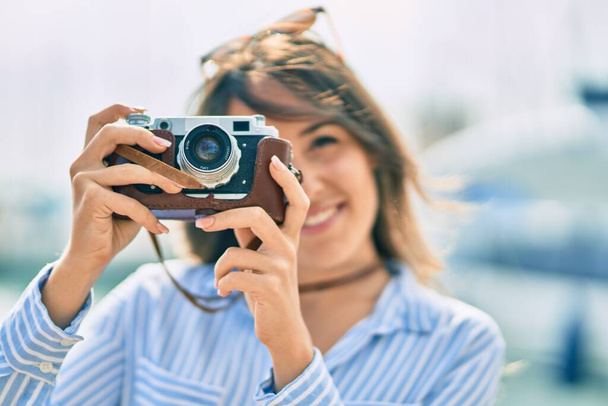 Jovem turista hispânica sorrindo feliz usando a câmera vintage no porto. - Foto, Imagem