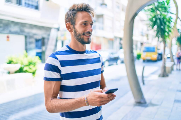 Όμορφος καυκάσιος άνδρας με γενειάδα χαμογελώντας χαρούμενος σε εξωτερικούς χώρους χρησιμοποιώντας smartphone - Φωτογραφία, εικόνα