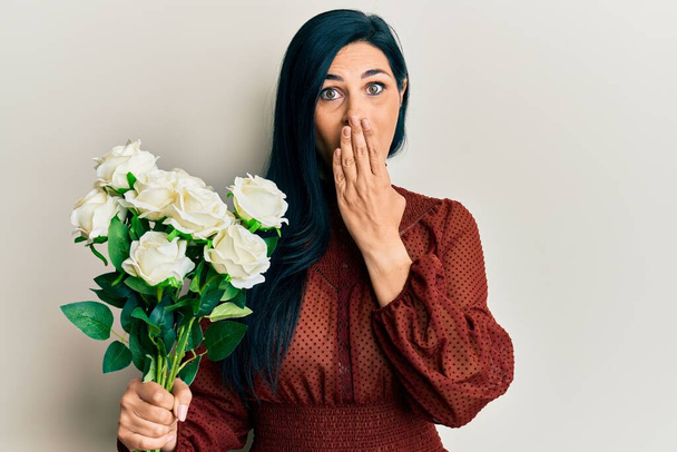 Young caucasian woman holding flowers covering mouth with hand, shocked and afraid for mistake. surprised expression  - Photo, Image