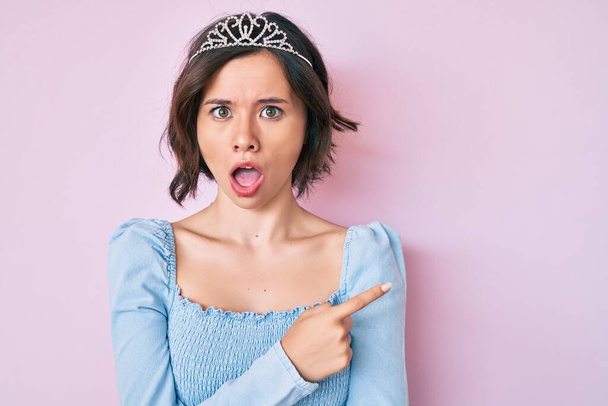 Young beautiful girl wearing princess crown in shock face, looking skeptical and sarcastic, surprised with open mouth  - Photo, Image