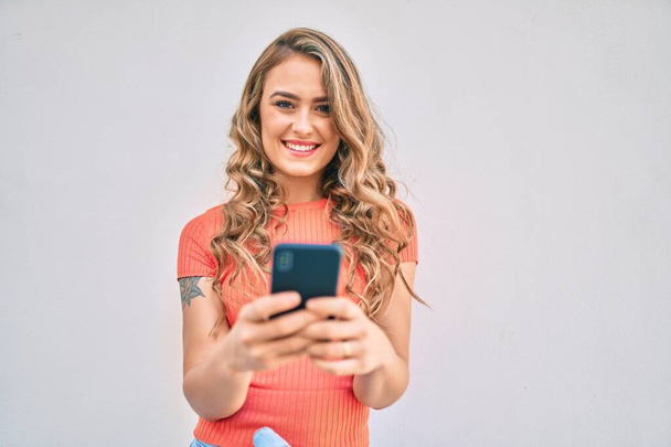 Jeune fille blonde souriant heureux en utilisant smartphone debout à la ville. - Photo, image