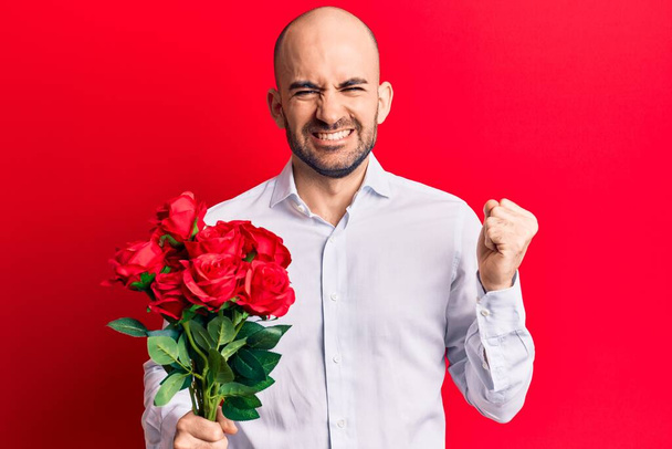 Genç ve kel adam elinde bir buket kırmızı çiçekle gururla bağırıyor, zaferi kutluyor ve kollarını kaldırdığında çok heyecanlanıyor.  - Fotoğraf, Görsel