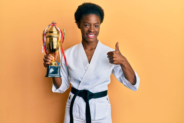 Giovane ragazza afroamericana indossa kimono karate tenendo trofeo sorridente felice e positivo, pollice in su facendo segno eccellente e approvazione  - Foto, immagini