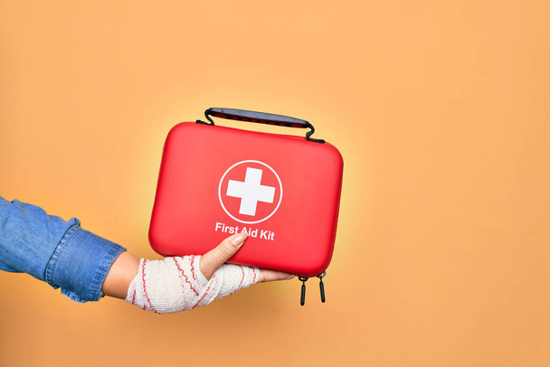 Wounded hand of caucasian young woman cover with bandage holding first aid kit briefcase over isolated yellow background - Zdjęcie, obraz
