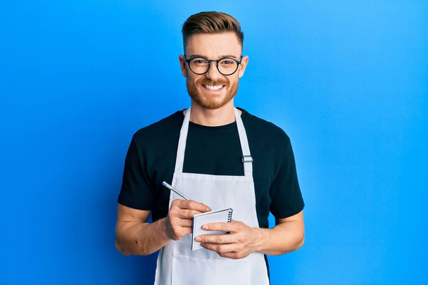 Young redhead man wearing waiter apron taking order smiling with a happy and cool smile on face. showing teeth.  - Foto, afbeelding