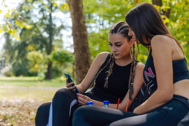 Beautiful young female friends sitting on a bench in a park while looking at a smartphone. - Foto, Imagem