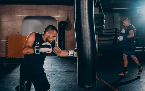 Férfi bokszoló edzés boxzsákkal a sötét sportcsarnokban. Férfi bokszoló, mint testmozgás a nagy harcra. Boxer találat boxzsák - Fotó, kép