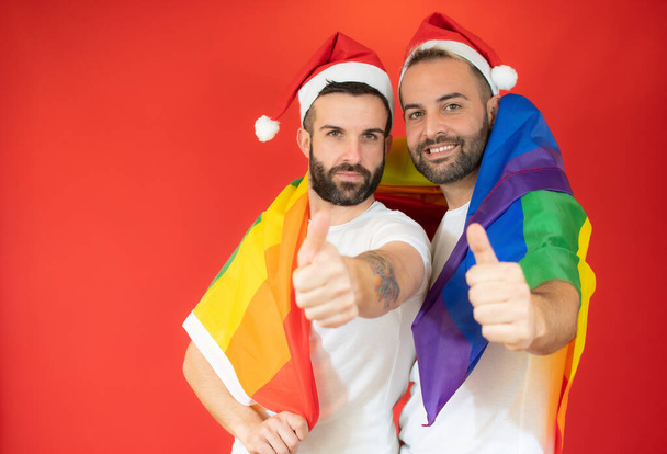 feliz pareja gay en santa hat y envuelto con la homosexual bandera con pulgares arriba sobre rojo fondo. - Foto, imagen