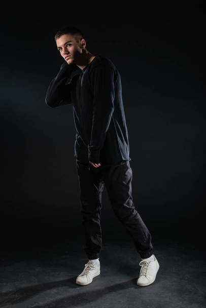 Ganzkörperbild eines attraktiven sexy jungen männlichen Modells, das isoliert auf schwarzem Hintergrund posiert und eine schwarze Bluse und schwarze Hose trägt. - Foto, Bild