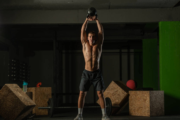 Full-length photo of a handsome man with a naked torso exercising with a kettlebell on a dark background - Foto, Imagem