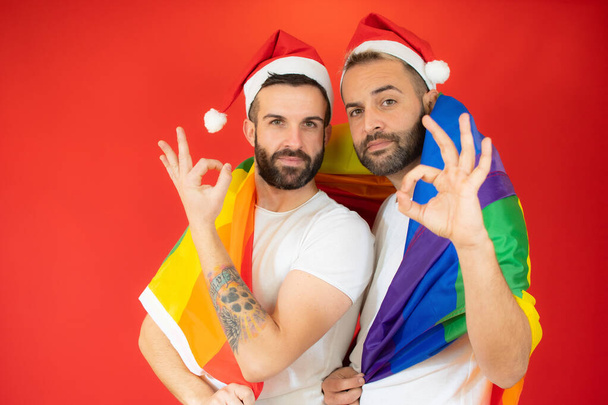 幸せなゲイのカップルでサンタの帽子と包まれています同性愛者の旗,赤の背景に大丈夫なサイン. - 写真・画像